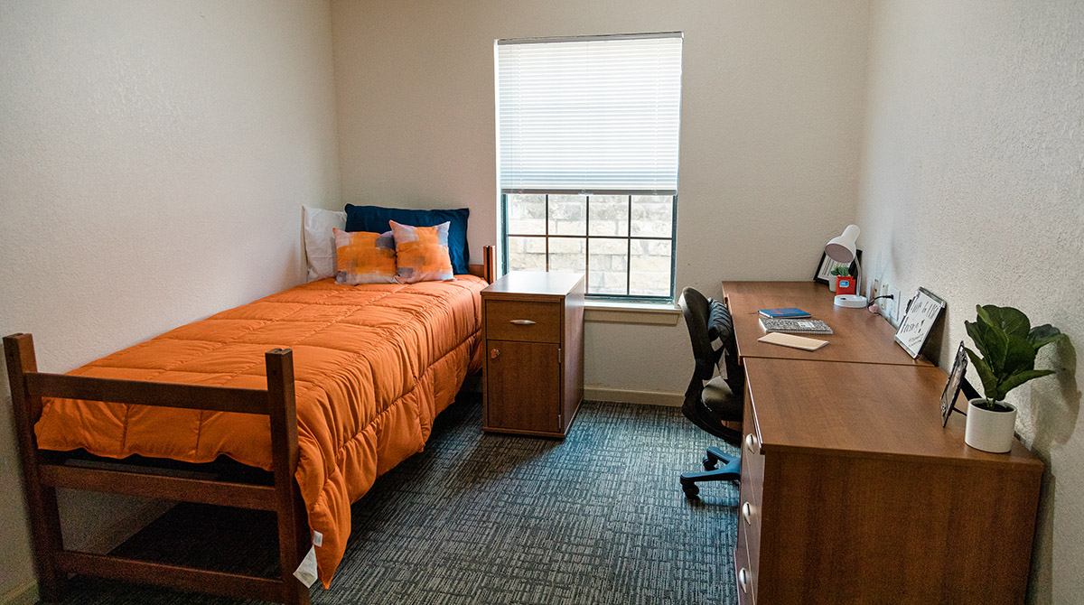 Photo of student room with bed, desk, closet, window, and nightstand.